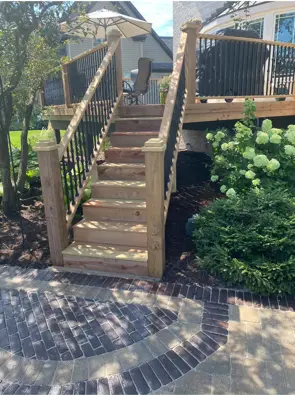 Wood deck stairs with black balusters beside a landscaped brick patio